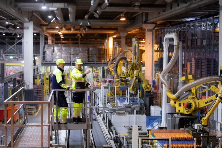 Kaksi henkilöä keltaisissa suojavaatteissa seisoo teollisuushallissa tarkastelemassa automaattisia tuotantolinjoja.