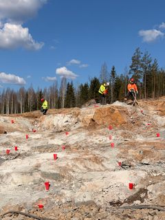 Kalliotyömaa, jossa useita porauspisteitä merkitty punaisin merkein ja työntekijät työskentelevät suojavarusteissa.