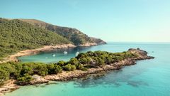 Näkymä Mallorcan rannikolta: turkoosi meri, vehreä mäntymetsä ja pieni poukama, jossa veneitä.