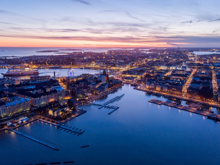 Ilmakuva kaupungista iltahämärässä, taustalla laskeva aurinko ja valaistuja katuja.