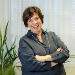 Nainen, tummat hiukset ja tummanharmaa paita, seisoo hymyillen vaalean verhon edessä.