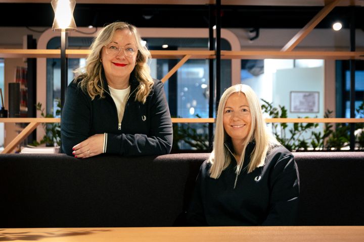 Kaksi naista toimistoympäristössä, mustat paidat. Iloiset, innokkaat ilmeet ja rento asento.