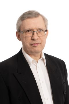 Misstänkt person, grått hår, glasögon, mörk kostym. Står och tittar på kameran.