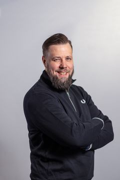 Mies, jolla tummat hiukset ja iloinen ilme. Katsoo kohti kameraa. Musta paita.