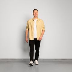 Mies katsoo kohti kameraa ja ottaa askeleen eteenpäin. Vaaleankeltainen paita, valkoinen t-paita, mustat farkut, valkoiset lenkkitossut.