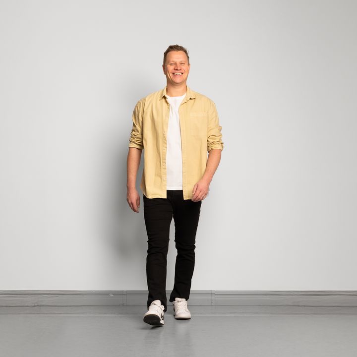 Mies katsoo kohti kameraa ja ottaa askeleen eteenpäin. Vaaleankeltainen paita, valkoinen t-paita, mustat farkut, valkoiset lenkkitossut.