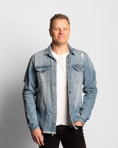 Vaalea mies, farkkupaita, valkoinen t-paita, mustat farkut. Seisoo ja katsoo kohti kameraa, iloinen ilme.