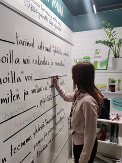 Nuori nainen kirjoittamassa jättikokoiselle valkotaululle messuilla.