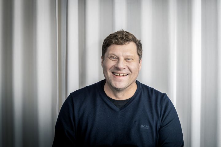 kuvassa mies mustassa neulepaidassa, katsoo ja hymyilee kameralle