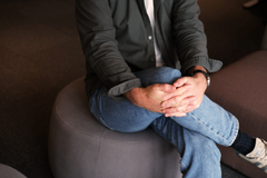 Henkilö istuu jakkaralla jalka toisen päällä ristissä, kädet ristissä jalkojen päällä.