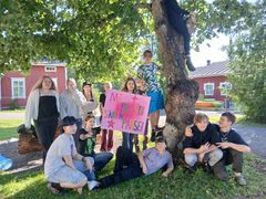 Osallistujia ryhmäkuvassa vanhan puun ympärillä, taustalla punainen rakennus.