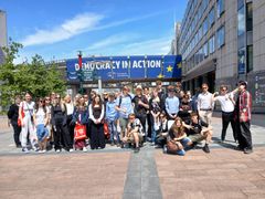 Ryhmä METÄS!-hankkeen osallistujia poseeraa ulkona Brysselissä. Taustalla näkyy rakennus, jossa lukee "Democracy in Action".