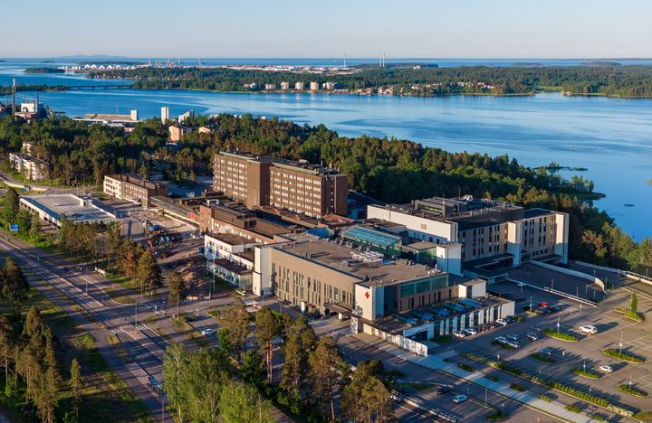 Ilmakuva sairaalarakennuskompleksista metsän ja veden läheisyydessä aurinkoisena päivänä.