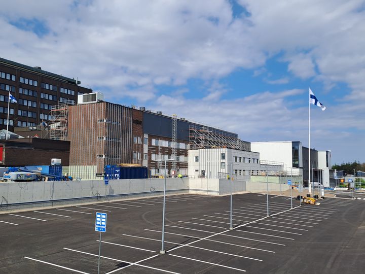 Rakennustyömaa Kymenlaakson keskussairaalan edessä, pysäköintialue edessä. Taustalla Suomen lippu liehuu lipputangossa.
