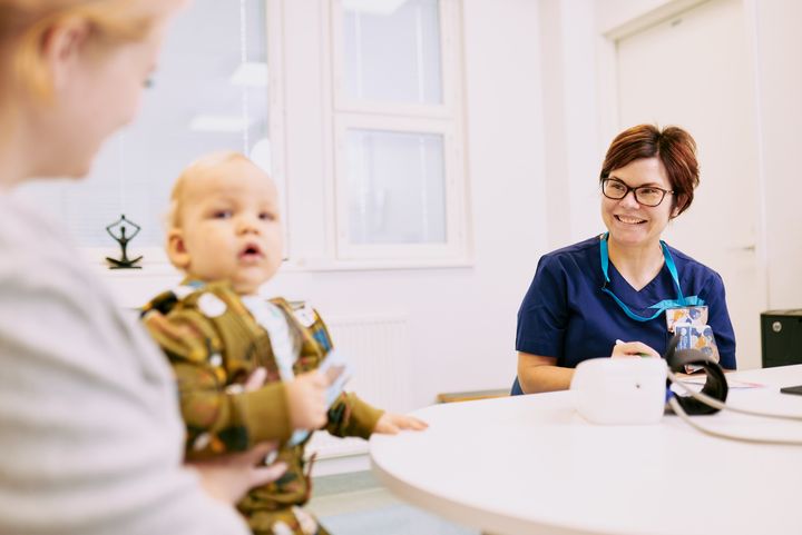 Aikuinen hymyilee hoitohuoneessa pitäen lasta sylissään, samalla kun toinen henkilö istuu ja hymyilee pöydän ääressä.