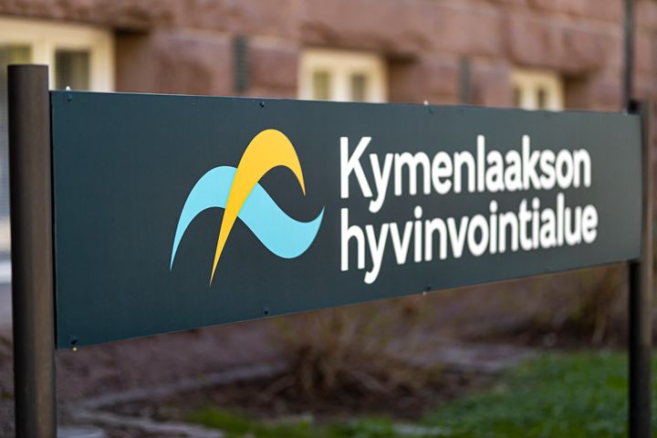 Kyltti, jossa lukee "Kymenlaakson hyvinvointialue" ja siinä on keltaisen ja sinisen kaaren muotoiset kuviot.