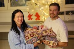 A family is smiling while holding two newborn babies wrapped in a crochet blanket, with holiday decorations in the background.