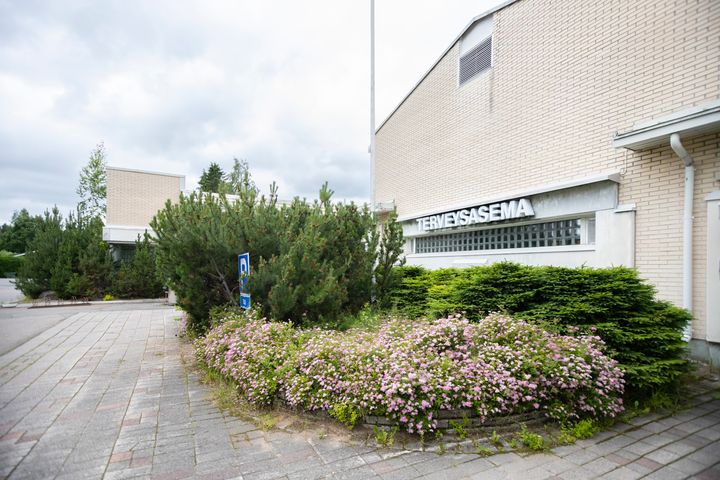 Rakennus, jossa lukee "terveysasema", ympäröitynä pensailla ja kukilla.
