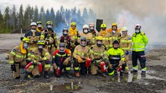 Ryhmä palomiehiä ja pelastushenkilöstöä poseeraa ulkona suojavarusteissa. Heillä on kypärät, suojapuvut ja jotkut pitävät palosammuttimia käsissään. Taustalla on savua ja metsää.