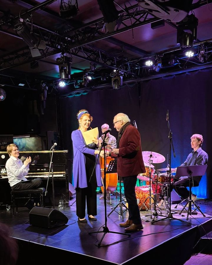 Neljä henkilöä esiintymislavalla. Kaksi keskellä olevaa kättelevät ja pitävät papereita. Yksi henkilö soittaa pianoa, toinen rumpuja. Taustalla tumma verho.