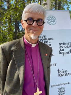 Piispa hymyilee kameralle Gobaali kirkko -julisteen edessä