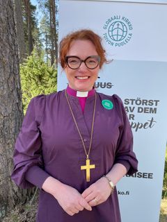 Piispa hymyilee kameralle Gobaali kirkko -julisteen edessä