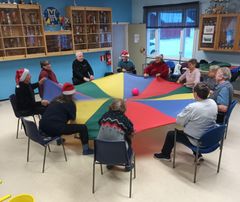 En grupp äldre och deras handledare sitter på stolar i en ring. De håller i kanterna på ett stort, runt, färgglatt tyg. Ovanpå duken finns en röd boll.