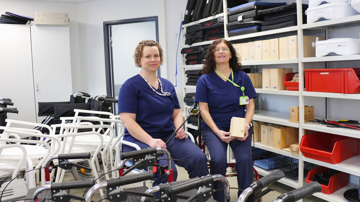 Två kvinnor med mörkblåa arbetskläder på sitter bakom rollator och håller hjälpmedel i handen.