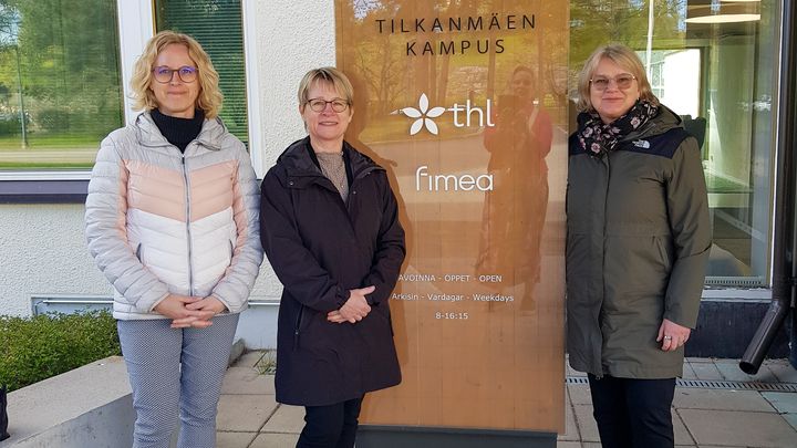 Kolme ihmistä seisoo vierekkäin rakennuksen sisäänkäynnillä.
