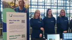 Kolme ihmistä seisoo pöydän takana marketin aulassa.