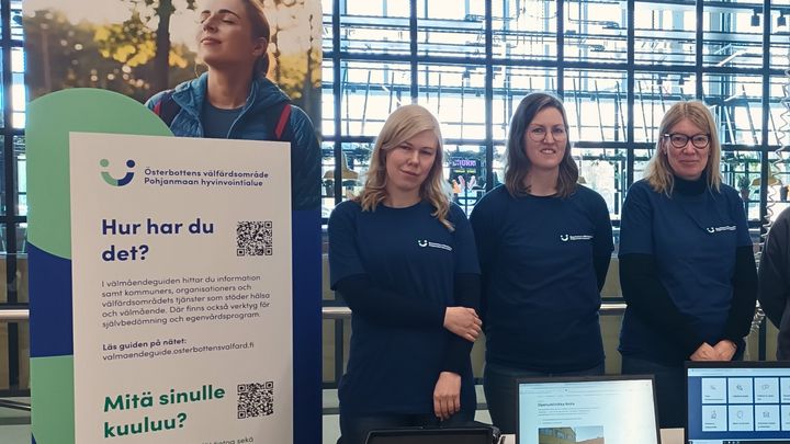 Kolme ihmistä seisoo pöydän takana marketin aulassa.