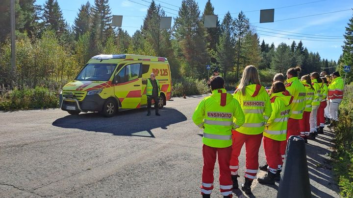 Ambulanssi parkkeerattuna ajoradalle, ambulanssin vieressä seisoo mies keltaisissa huomioliiveissä. Rivi ensihoitajia puna-keltaisissa työvaatteissaan katsoo ambulanssin suuntaan.