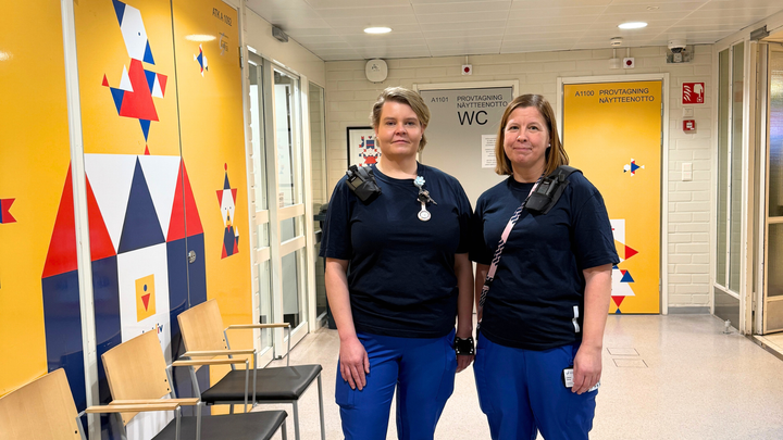 Två kvinnor, klädd i sjukskötarens arbetskläder står i korridor. Korridorens vägg är färgfull.