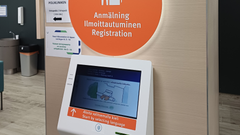 På bildens övre kant syns en stor orange skylt på väggen, där det står på finska, svenska och engelska att anmäla sig. Under skylten finns en skärm för anmälningsautomaten.