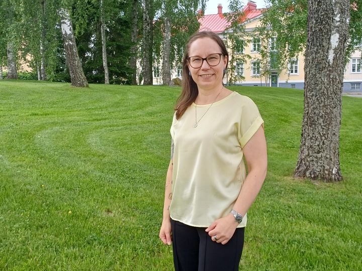 Projektchefen Sonja Kotala ler mot kameran. Hon har en gul tröja och står bredvid en björk utomhus på sommaren.