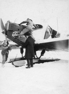 Alt-teksti: Mustavalkoisessa valokuvassa vuodelta 1926 on vanha lentokone, jota mekaanikko pyörittää käynnistääkseen sen. Siivellä seisoo lentäjä talvivaatteissa, kun taas toinen henkilö seuraa tilannetta etualalla. Taustalla näkyy lumipeitteinen maisema.