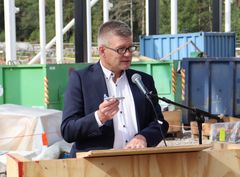 Mies puhumassa mikrofonin edessä rakennustyömaalla, pitäen pienoismallia käsissään.