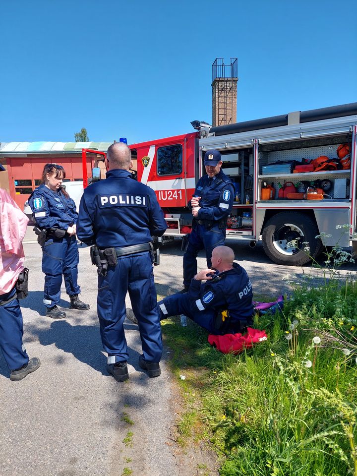 Poliisiasuisia%20taustan%E4yttelij%F6it%E4%20istuu%20vilvoittelemassa%20paloauton%20varjossa%20kuvauspaikalla.