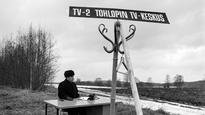 Mies istumassa toimistopöydön ääressä peltomaisemassa. Naulakkoon on lisätty kyltti, jossa lukee TV2, Tohlopin tv-keskus.
