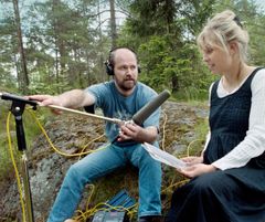 Metsäisessä puistossa ison kiven päällä istuu ääniteknikkomies, jolla on edessään iso mikrofoni. Vieressä on kirjailija Laura Ruohonen, jolla on käsissään luettavaa.