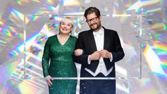 Kaksi henkilöä hymyilee ja poseeraa yhdessä juhlapuvuissa värikkäässä taustassa.