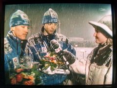 Hiihtäjät Isometsä ja Myllylä talvivaatteissa pitävät kukkakimppuja lumisateessa, toimittaja ojentaa mikrofonia.