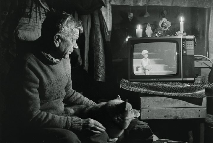 Vanha mies katsoo televisiota hämärässä huoneessa, koira sylissään. Televisiossa näkyy urheilulähetys.