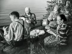 Neljä henkilöä syö eväitä kallion päällä järven rannalla ja kuuntelee radiota.