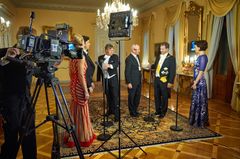 Ryhmä ihmisiä juhlapuvuissa keskustelee suuressa, koristellussa huoneessa. Taustalla on valokuvauskalustoa.