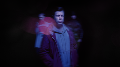 Mies seisoo tummassa ympäristössä, yllään violetti takki ja huppari. Taustalla on kaksi hahmoa epätarkasti.