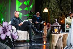 Kolme henkilöä istuu lavalla, lukevat kirjoja. Lavastus sisältää kasvikuvioita ja lamppuja.