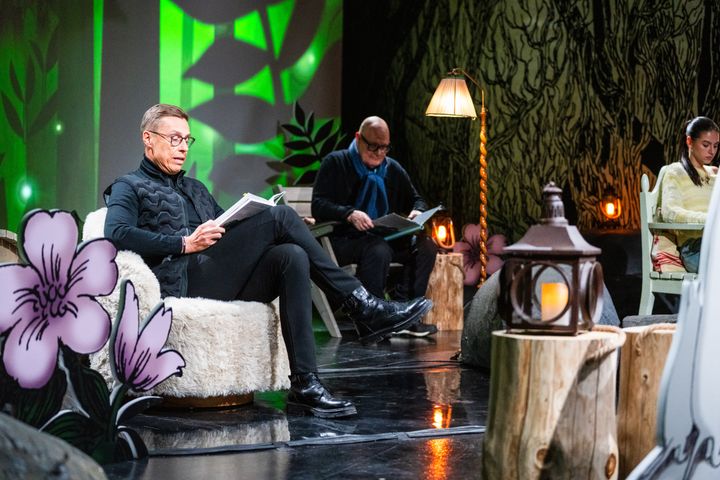Kolme henkilöä istuu lavalla, lukevat kirjoja. Lavastus sisältää kasvikuvioita ja lamppuja.