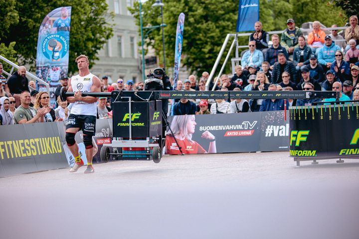 Suomen Vahvin Mies (105 kg) -finaalin 2026 lajit ovat tukkipunnerrus, maastaveto, etupito, ski erg, hiekkasäkkiestejuoksu, ruorihinaus, Herkuleen pito, merimieskävely, säkinheitto ja atlaskivet. Kuva vuoden 2025 finaalista ja Tino Näkkilästä, lajina conanpyörä.
