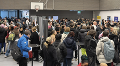 Sisätiloissa järjestetty tapahtuma, jossa suuri joukko ihmisiä seisoo tiiviisti esittelypisteiden ja roll-upien ympärillä; osallistujilla on talvivaatteet ja reppuja, ja tila on liikuntasali.